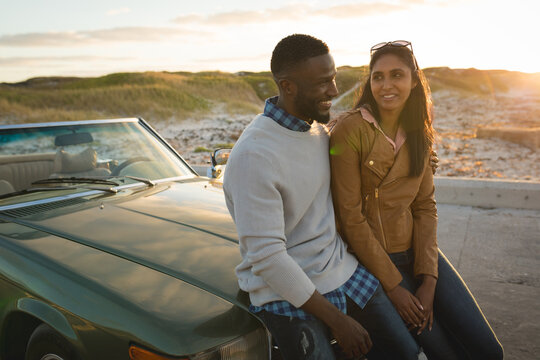 Diverse couple sitting on a convertible car looking at each other and smiling