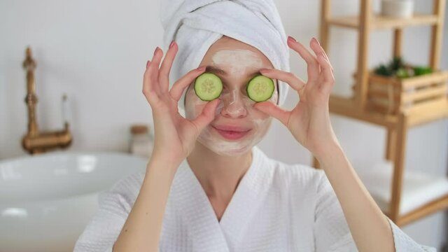 Skin Care, After Bath, Cosmetics at Home, Caucasian Woman, Anti Aging. Cheerful caucasian woman in a robe and a towel on her head uses a cosmetic face mask and cucumber