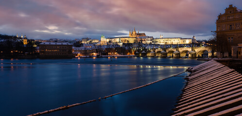 panoramic view of the snow-covered Prague Castle and St. Vitus Cathedral and Charles Bridge on the Vltava River and on the banks of a wooden barrier and colorful sky
