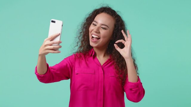 Cheerful african american woman 20s in pink shirt isolated on blue background studio. People lifestyle concept. Doing selfie shot on mobile phone showing victory sign ok okay blowing sending air kiss