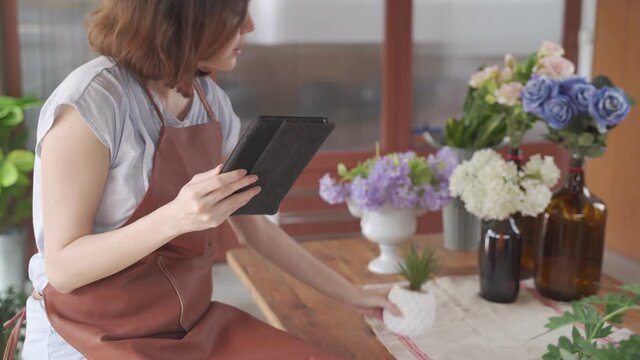 Young asian female entrepreneur touching tablet screen searching information looking at flowers in florist's shop enjoying nature and modern technology. People, entrepreneurship and business concept.