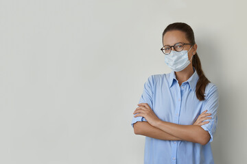 Young woman in blue shirt with arms crossed looking at the camera, wearing protective face mask and glasses
