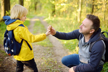 Fototapeta na wymiar Cute schoolchild and his mature father hiking together and exploring nature. Little boy with his dad spend quality family time together in the sunny summer forest.