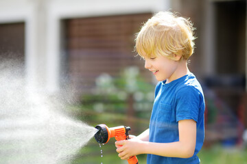 Funny little boy watering lawn and playing with garden hose with sprinkler in sunny backyard....