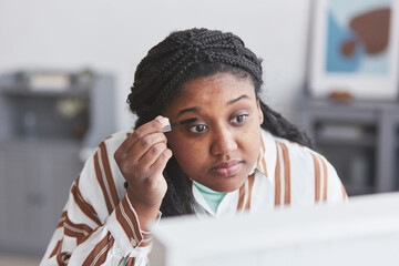 Close up portrait of curvy African-American woman putting on makeup and looking in mirror while getting ready to go out at home, copy space