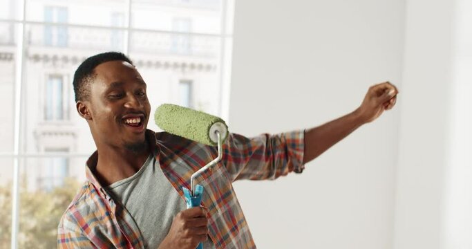 Portrait of young african american cheerful man having fun in new apartment making repairs dancing and singing using paint roller as microphone.