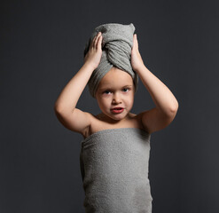 pretty girl of preschool age, after a shower, wrapped in a gray terry towel, holds a rolled towel on her head with both hands.