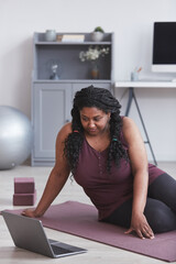 Vertical portrait of curvy African American woman working out at home while sitting on yoga mat and...