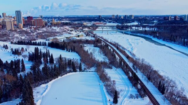 Aerial flyover winters edge condos apts by cliff of Victoria Park manmade quiet ice rink connected to an infinity skate track next to forest road snow covered North Saskatchewan River downtown city2-4