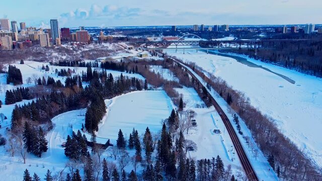 Aerial flyover winter Victoria Park manmade quiet ice rink connected to an infinity skate track next to forest road next to snow covered icy North Saskatchewan River edge of condos downtown city1-4