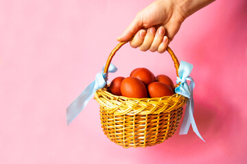 Womans hand holds a wicker basket decorated by blue ribbons with  brown Easter eggs on a pink...
