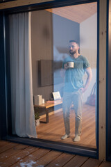 Young bearded man holding mug with hot drink and looking through large window
