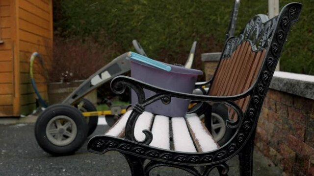 Snow falls on a cast iron chair in the middle of winter. A washing bucket and kids toys can be seen in the background. Snow flakes swirl around in the strong wind. A typical scene of an Irish winter.