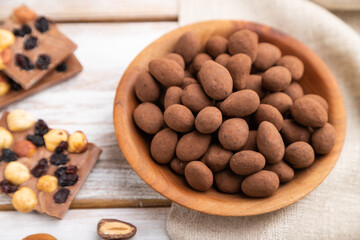 Almond in chocolate dragees in wooden plate and a cup of coffee on white wooden background. Side view, selective focus.