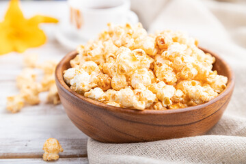 Popcorn with caramel in wooden bowl and a cup of coffee on a white wooden background. Side view, selective focus.