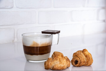 Glass Cup of tricolour Cappuccino, with chocolate croissant, and piece of chocolate. Wooden wall background and white floor.