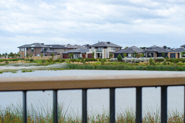 View of a row of modern suburban houses by a lake over the fence of a viewing platform. Concept of real estate development, the housing market, Australian homes, and natural environment in new suburb.