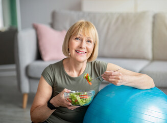 Exercise and healthy diet concept. Senior woman with fitball eating fresh vegetable salad at home - obrazy, fototapety, plakaty