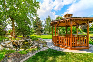 A nicely landscaped garden and back yard with a round wooden cedar gazebo and a pond with waterfall - obrazy, fototapety, plakaty