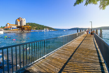 Fototapeta na wymiar A young boy leans over the dock edge at afternoon at a lakefront wooden dock with a hotel resort and mountain behind in Coeur d'Alene Idaho USA 