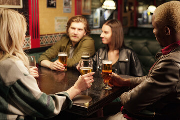 Young intercultural friends sitting by table in pub, having beer and discussion