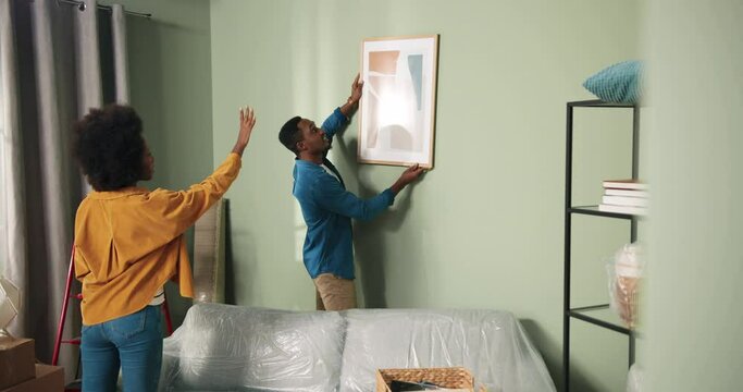 Happy African American couple has moving into new apartment and are decorating space. Stylish girl telling guy how to hang picture.