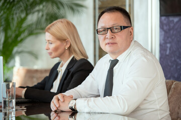 Mature business delegate sitting by table against his elegant blond secretary