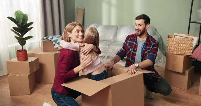 Young happy caucasian couple opens box with daughter in new apartment. Little girl jumps out of cardboard box and hugs her parents.