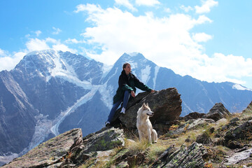A young woman tourist climbed the mountain with her white husky dog. They sit on a rock and admire the mountain panorama. Inseparable owner and pet. Active lifestyle. Travel through the countryside