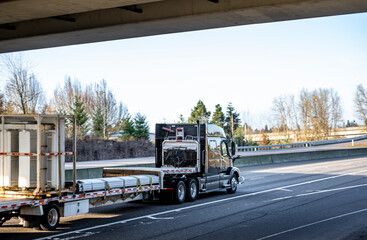 Black big rig semi truck with low cab transporting industrial cargo on flat bed semi trailer running on the road under the bridge