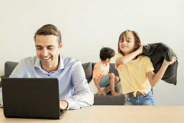 Cheerful dad chatting via laptop and kids playing with pillows near him. Caucasian father working at home during school vacations. Happy children on background. Family and digital technology concept