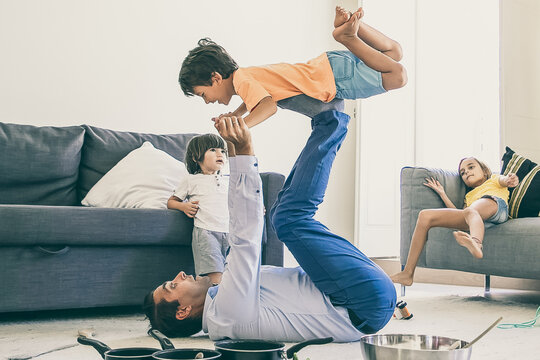 Caucasian dad holding son on legs and lying on carpet. Happy cute boy flying in living room with help of father. Cute kids playing together near bowl and pans. Childhood and weekend concept