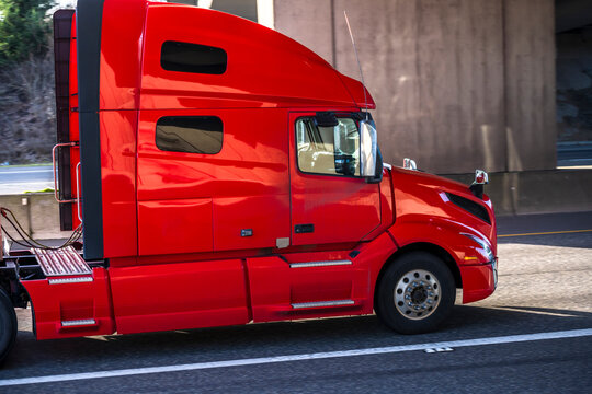 Bright red gorgeous big rig semi truck tractor transporting cargo running on the divided highway road going under the bridge