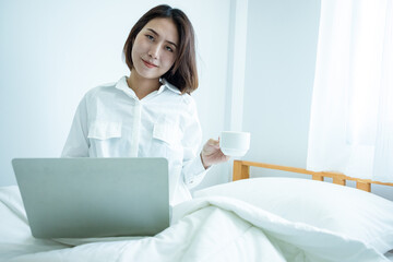 Happy casual beautiful Asian woman working on a laptop laying and drinks coffee on the bed in the house after wake up. Working from home in quarantine lockdown.