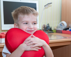 Adorable boy holding a heart-shaped pillow in his hands at home. Happy birthday greeting, Happy Valentine's Day concept