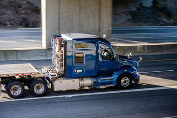 Shiny blue big rig semi truck with long cab and flat bed semi trailer driving on the highway road under the bridge
