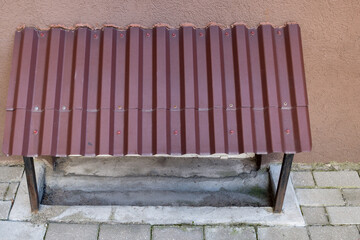 A maroon corrugated metal sunshade visor attached to iron posts embedded in the cement floor above the basement window against a background of brown wall and area of paved gray street tiles.