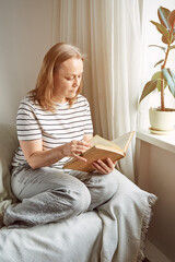 A mature woman in home clothes, sitting on the couch, reads a book near the window.