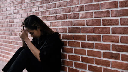 Christian Asian woman is sitting against a wall, feeling sad, holding a cross and praying to God.