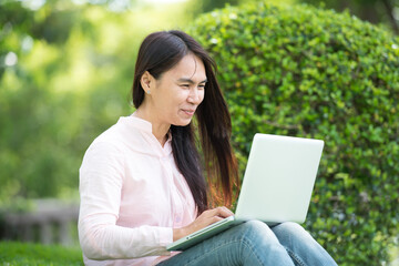 Asian woman sitting green park using laptop computer. Woman working on laptop happy entrepreneur business using notebook with hands typing on keyboard home office during coronavirus quarantine period