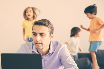 Serious Caucasian dad working via laptop and kids jumping on background. Concentrated father using computer and kids playing on sofa. Selective focus. Childhood and digital technology concept
