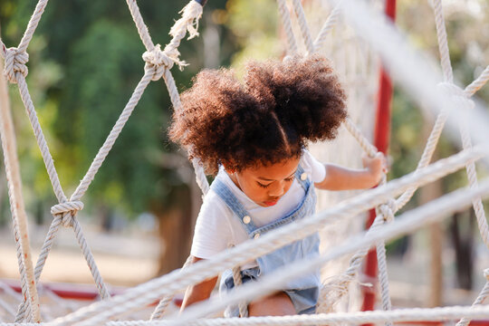 Little African American curly hair girl playing on park playground in spring or summer season. They enjoy climbing on the rope equipment.