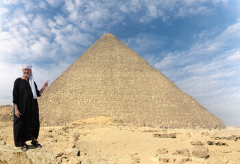 An Egyptian in a black robe and a turban stands in front of the huge Cheops pyramid in Giza. He...