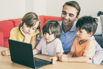 Cheerful dad and pensive kids watching movie via laptop together during weekend. Happy father sitting at table with children in living room. Fatherhood, childhood and digital technology concept