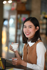 Portrait of cheerful young female holding a cup of coffee and smiling to camera while sitting in modern coffee shop.