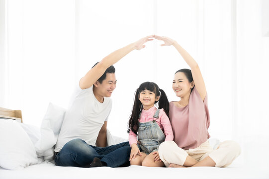 Happy Asian Family Sitting On Bed In Bedroom Together And Making The Home Sign. Mom And Dad Making Roof Figure With Hands Arms Over Heads