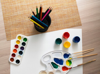 a blank sheet of white paper, a palette, watercolors and acrylics, brushes, a stand with colored pencils on a light background