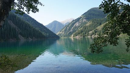 Kolsay lake among green hills and mountains. The mountain lake is surrounded by green forest, tall coniferous trees, grass and bushes. Clean water is like a mirror. Tourists swim on boats. Kazakhstan - Powered by Adobe