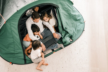 Top-view of happy kids relaxing with father in tent at home and watching cartoon on laptop...