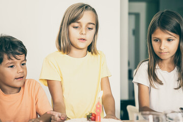 Cute blonde girl making wish and celebrating her birthday with friends. Lovely children standing in room together and looking at tasty cake with candle. Childhood, celebration and holiday concept